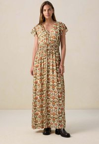 Maxi-jurk in crème met een bloemenprint in warme kleuren. Heeft een V-hals, korte geplooide mouwen en een elastische tailleband. Gemaakt met zwarte laarzen.