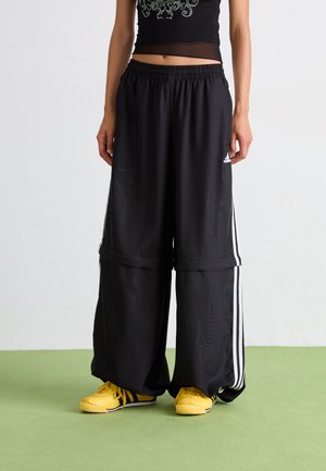 Pantalons de sport noirs à jambes larges avec une taille élastique et des rayures blanches sur les côtés, associés à des baskets jaunes.