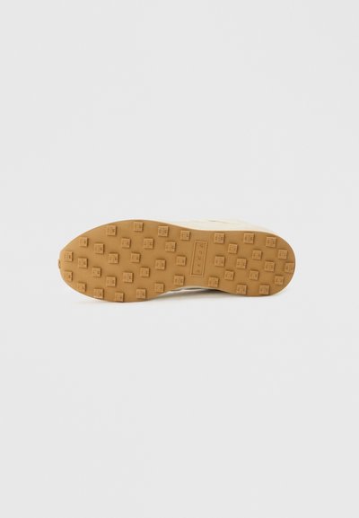 Μπεζ σόλα αθλητικού παπουτσιού με τετράγωνα ανάγλυφα πέλματα και το όνομα της μάρκας χαραγμένο στο κέντρο σε ανοιχτό γκρι φόντο.