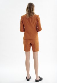 Oranžový přizpůsobený blejzr v kombinaci s odpovídajícími šortkami. Hladká textura, strukturovaný design a minimální hardware. Černé slip-on boty.