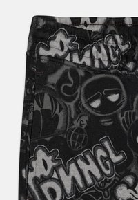 Pantalons en tissu extensible noir avec un imprimé graphique présentant divers dessins et textes, mettant en avant un mélange de textures et de motifs.