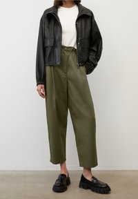 Persona che indossa una giacca di pelle nera corta, una t-shirt bianca, pantaloni a vita alta verde oliva a gamba larga e mocassini neri spessi.