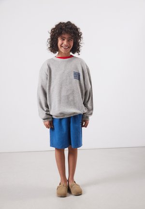 Smiling child with curly hair wearing gray sweatshirt, blue shorts, and tan slip-on shoes standing against plain background.