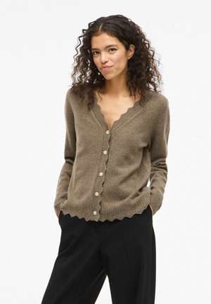 Cardigan marrone con bottoni, dal colletto e dall'orlo ondulati, texture morbida, con bottoni chiari e una vestibilità rilassata. Pantaloni neri sottostanti.
