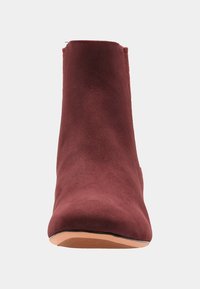 Przód bordowego, zamszowego botka z zaokrąglonym noskiem i beżową gumową podeszwą na jasnym tle.