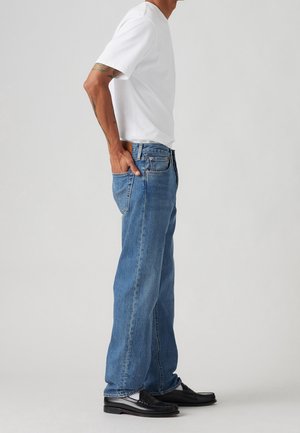 Person iført en simpel hvid t-shirt, blå straight-leg jeans og sorte loafers, stående i sideprofil med den ene hånd i baglommen.