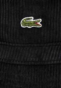 Cappello a secchiello in corduroy nero con un piccolo logo di coccodrillo ricamato verde con accenti rossi sulla parte anteriore. Tessuto morbido e testurizzato con coste.