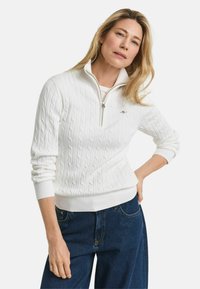 Weißer Pullover mit Kabelmuster und Reißverschluss im Stehkragen, mit Rippbündchen und -saum, kombiniert mit dunkelblauen weiten Jeans. Einfache Hintergrund.