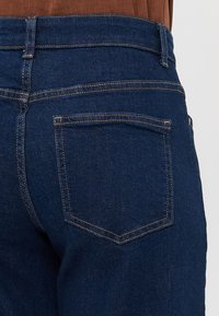 Jeans de ganga azul escuro com um silhueta ajustada, apresentando dois bolsos traseiros e detalhes de costura visíveis ao longo das costuras.