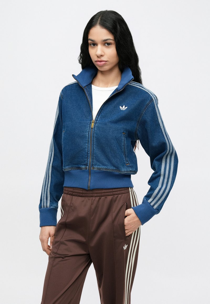 Chaqueta vaquera corta en azul oscuro con cuello alto, cremallera dorada y detalles a rayas grises. Llevada sobre un top blanco con pantalones deportivos marrones.