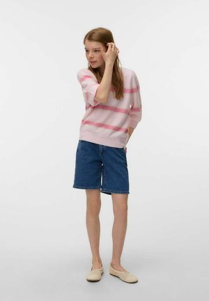 Jeune femme portant un pull rayé rose, un short en jean et des chaussures beige slip-on, debout et ajustant ses cheveux sur un fond blanc.