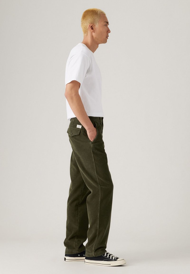 Khaki green corduroy pants with a relaxed fit, side pockets, and a white tag on the back. Worn with a plain white t-shirt and black sneakers.