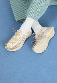 Des chaussures beiges à enfiler avec un dessus tissé, présentant une semelle texturée et des détails en dentelle. Portées avec des chaussettes blanches et un pantalon rayé vert sur une surface bleue.