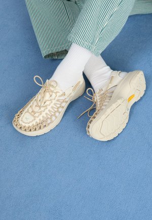Scarpe beige slip-on con materiale superiore tessuto, dotate di una suola testurizzata e dettaglio di lacci. Indossate con calze bianche e pantaloni a righe verdi su una superficie blu.