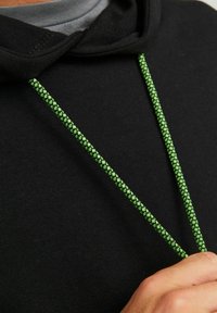 Un sweat à capuche noir avec des cordons tressés verts contrastants ; la vue en gros plan met en valeur la texture du matériau et les détails du design.