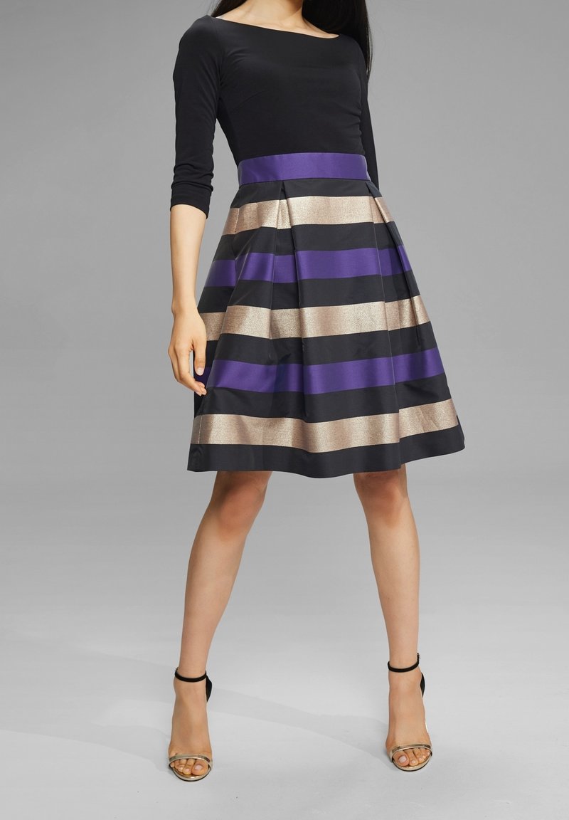 Femme portant une robe noire à manches coudées et une jupe plissée avec des rayures horizontales violettes, dorées et noires, associée à des talons noirs.