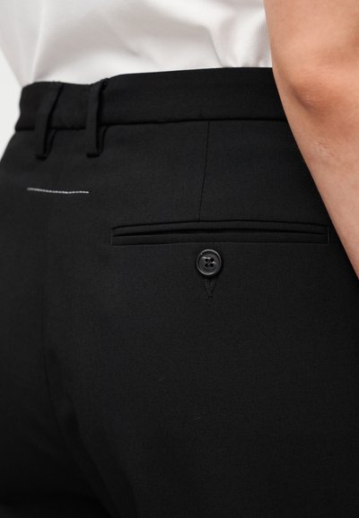 Pantalon noir avec une texture lisse, une poche arrière avec un bouton noir, et un détail de couture blanche subtile près de la ceinture.