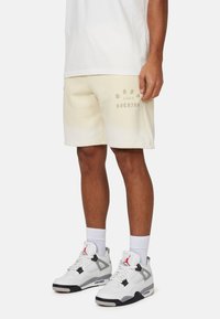 Pantaloni corti in cotone di colore crema con testo grafico in verde chiaro. Abbinati a sneakers bianche e grigie, con dettagli in nero e rosso.