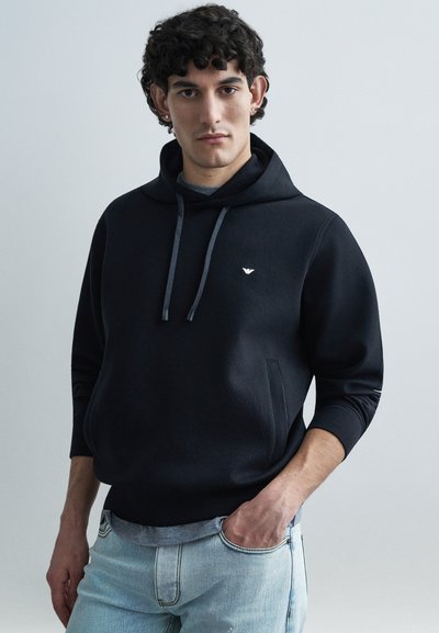 Sudadera negra con capucha, bolsillo frontal y cordones, con un pequeño logo en el pecho. Combinada con jeans azul claro. Textura de tela suave.