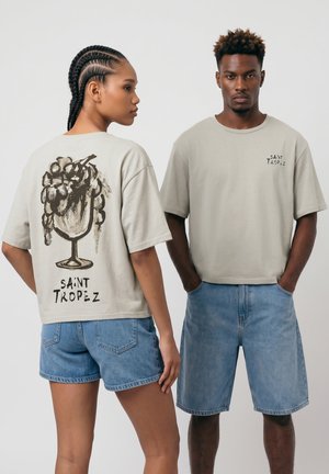 Dwie modelki noszą beżowe koszulki Saint Tropez i niebieskie krótkie spodenki jeansowe, jedna pokazuje przód, a druga tył z grafiką kieliszka z winogronami.