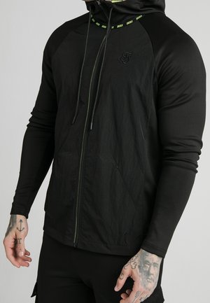 Haut à zip noir avec un tissu mat et texturé contrastant, cordons de serrage verts et accent logo sur la poitrine. Manches longues et poches.