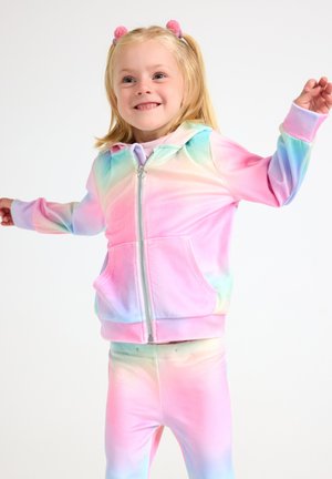 Młoda dziewczyna uśmiechająca się z wyciągniętymi ramionami, ubrana w pastelową bluzę z kapturem na zamek i dopasowane spodnie w stylu tie-dye na białym tle.