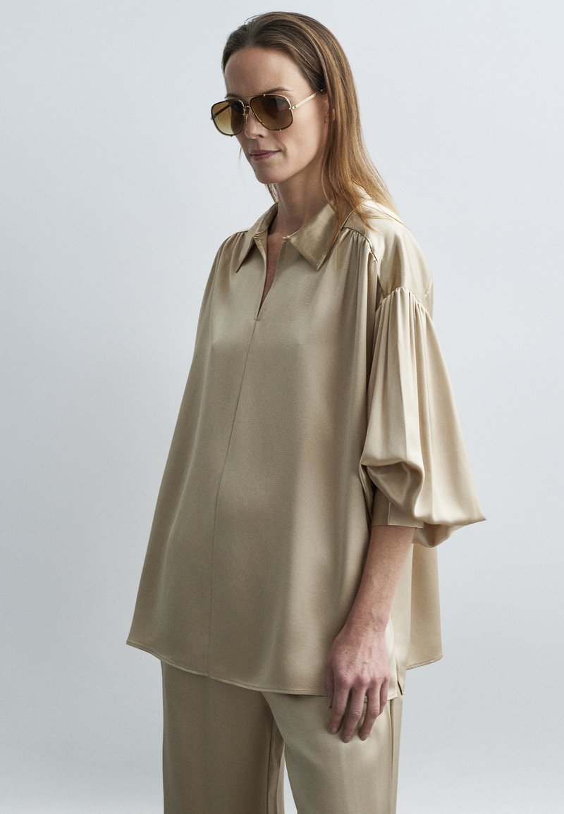 Žena v nadrozmernej béžovej saténovej blúzke so záhybmi na rukávoch a zladených nohaviciach, s veľkými hnedo sfarbenými slnečnými okuliarmi, na jednoduchom sivom pozadí.