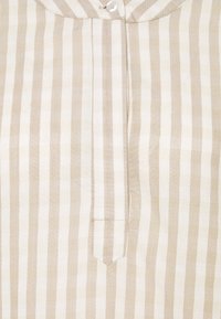 Lätta, beige och vita randiga tyg. Har en knapp-down krage och en vertikal placket med en liten ficka på sidan.