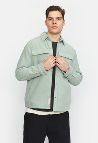 Lichtgroene corduroy shirt met een knopenfront, twee borstzakken en een puntige kraag, gelaagd over een zwart T-shirt en een wit ondershirt.