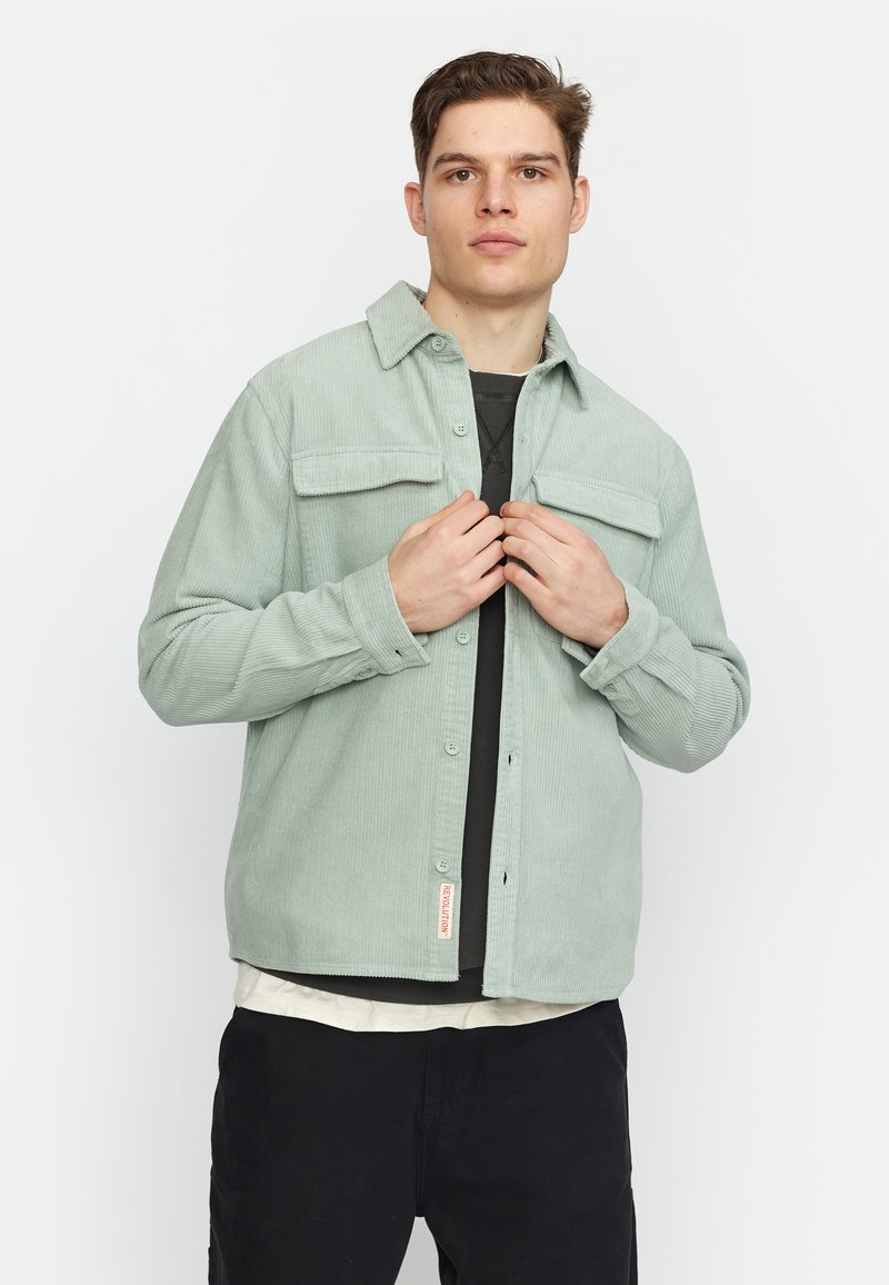 Lichtgroene corduroy shirt met een knopenfront, twee borstzakken en een puntige kraag, gelaagd over een zwart T-shirt en een wit ondershirt.