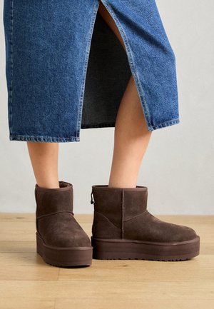 Bottes compensées en daim marron portées avec une jupe midi en denim bleu fendue à l'avant au-dessus des chevilles, sur un sol en bois.