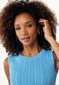 Haut sans manches bleu clair avec une texture côtelée et un col festonné. Accentué par un délicat collier en or avec un petit pendentif noir.