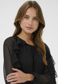 Ung kvinde med lysebrunt hår iført sølvfarvede hoopsøreringe og en sort gennemsigtig bluse med flæser på ærmerne, med armene over kors mod en hvid baggrund.