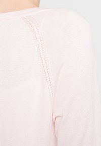 Světle růžový svetr s hladkou texturou, kulatým výstřihem a jemnými švy, které jsou doplněny drobnými výřezy na rameni.