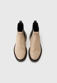 Cizme din suede bej până la glezne, cu laterale elastice negre, vârf rotund și o talpă texturată de culoare maro. Dispun de un mic accent din piele la toc.