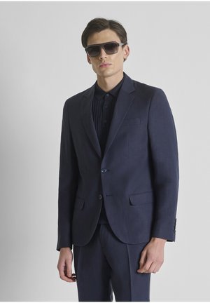 Man in een donkerblauw pak met dichtgeknoopte jas, zwart overhemd en grote donkere zonnebril, staand tegen een effen lichte achtergrond.