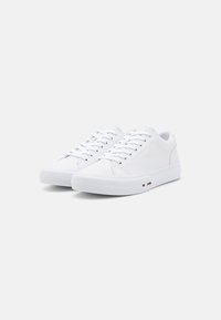 White canvas sneakers with a rubber sole, round toe, and flat laces. Features subtle branding on the side in red, white, and blue accents.