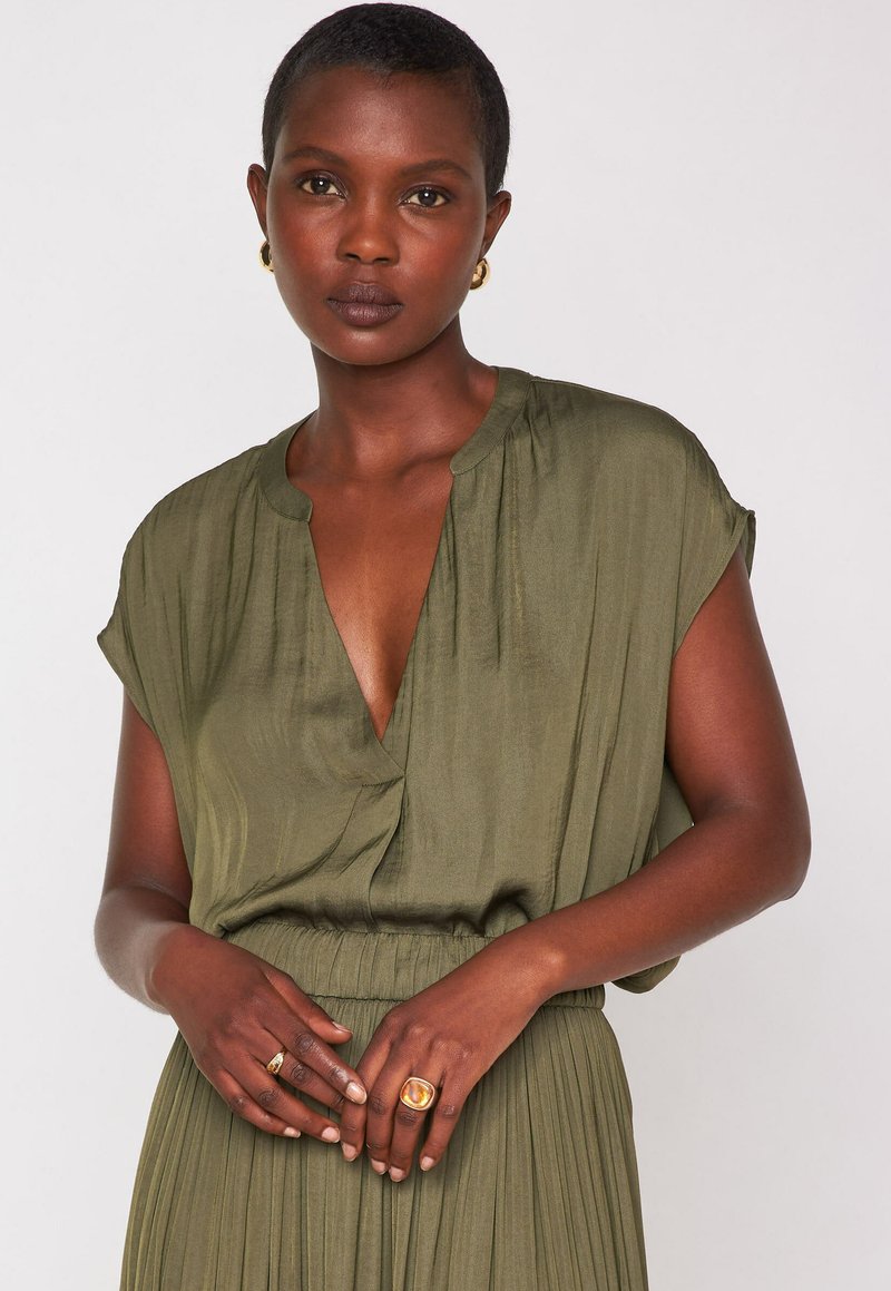 Vestito verde oliva a maniche corte con scollatura a V, texture plissettata e vita stretta. Anelli dorati sulle mani; finitura in tessuto liscia.