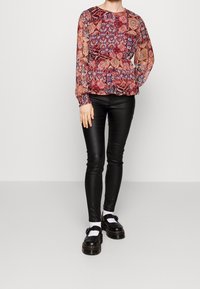 Camicia trasparente con motivo nei toni rosso, blu e beige, abbinata a pantaloni in pelle neri e aderenti e scarpe massicce nere con calzini bianchi.