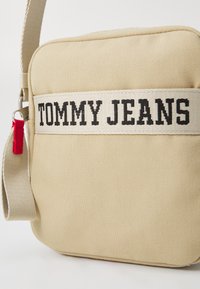Beige lerret crossover-veske med et vevd "TOMMY JEANS" merke i svart, med en rød glidelås og en avtagbar stropp.