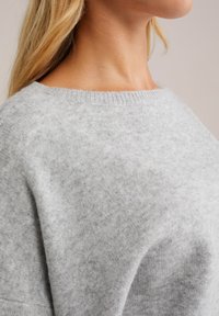 Pulover gri deschis, fabricat dintr-un material moale și texturat. Are un guler lat cu model în dungi și o croială lejeră, cu linii curate și detalii minime.