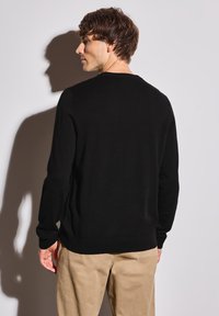 Pull en maille noire avec un col rond et des poignets côtelés, arborant une texture lisse. Associé à un pantalon de couleur claire, montrant un dos ajusté.