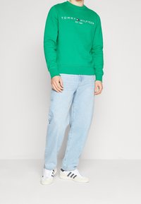 Zelená mikina s bílým logem "TOMMY HILFIGER", spárovaná s světle modrými volnými džíny a bílými teniskami s černými pruhy.