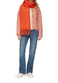 Veste à motif pied-de-poule en rose et beige, écharpe orange avec franges, pull léger, jeans évasés bleus et baskets blanches.