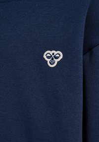 Navyblauer Stoff mit kleinem weißem besticktem Bienenlogo, mittig im Brustbereich eines Kleidungsstücks.