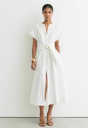 Vrouw draagt een witte midi-jurk met een brede riem en gouden gesp, een gouden armband en gouden open-toe sandalen, staand tegen een witte muur.