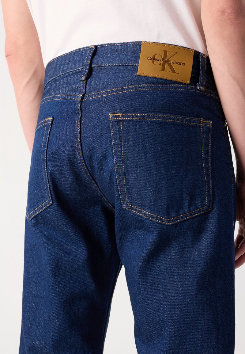 Granatowe dżinsy o gładkiej fakturze, z skórzaną metką z logo w talii oraz standardowymi tylnymi kieszeniami z żółtymi przeszyciami.