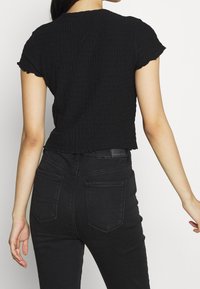 Young woman wearing a black textured short-sleeve top and high-waisted black jeans, shown from the back against a plain white background.