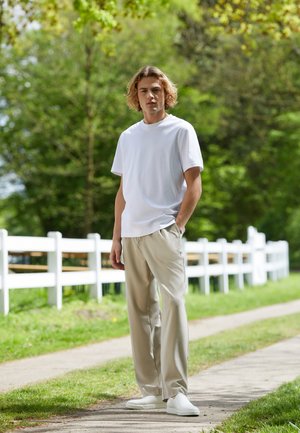 White cotton t-shirt, beige trousers, and white slip-on shoes. Model stands on a pathway with greenery and a white fence in background.
