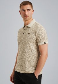 Polo beige con un motivo testurizzato di linee marroni, caratterizzato da un colletto a contrasto e un piccolo logo sul petto. Maniche corte.